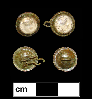 Copper alloy button back with a dark black or purple glass inset, flat on the front and beveled at the edge. The copper corrosion of the button back is toxic to microorganisms, which likely explains how some fabric that was in contact with the button survived and is still attached to the button back. The weave is visible, and the fabric is probably linen.